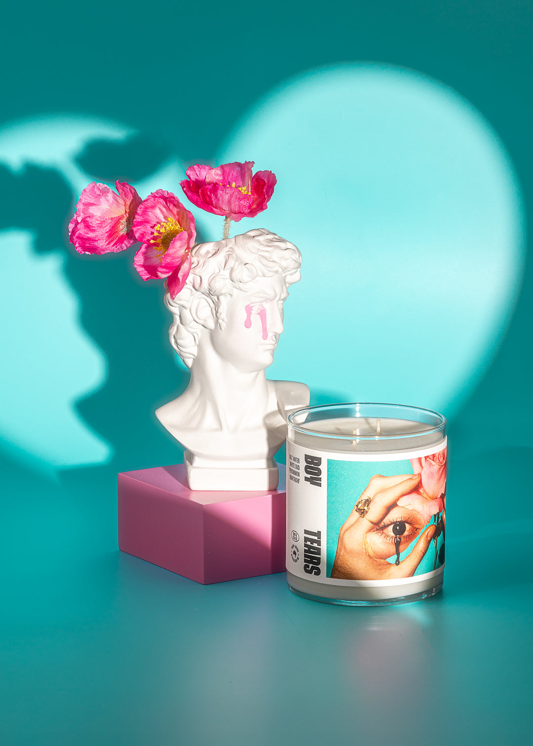 Candle with artistic design on a teal background with a bust and flowers.